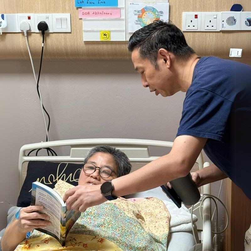Person reading a book in a hospital bed while another person assists nearby.